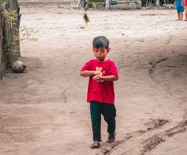 A young boy in red shirt and black pants standing on dirt.